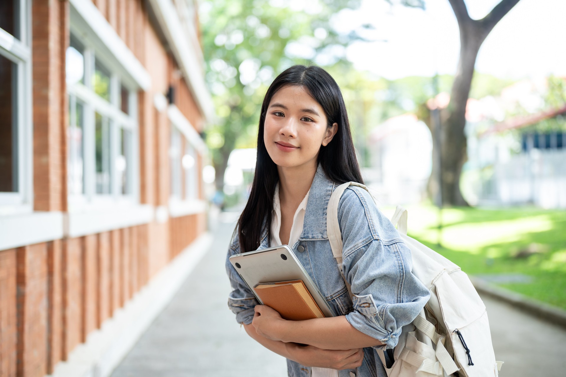 Young asian woman student with backpack holding hugging book and laptop on sidewalk around building.